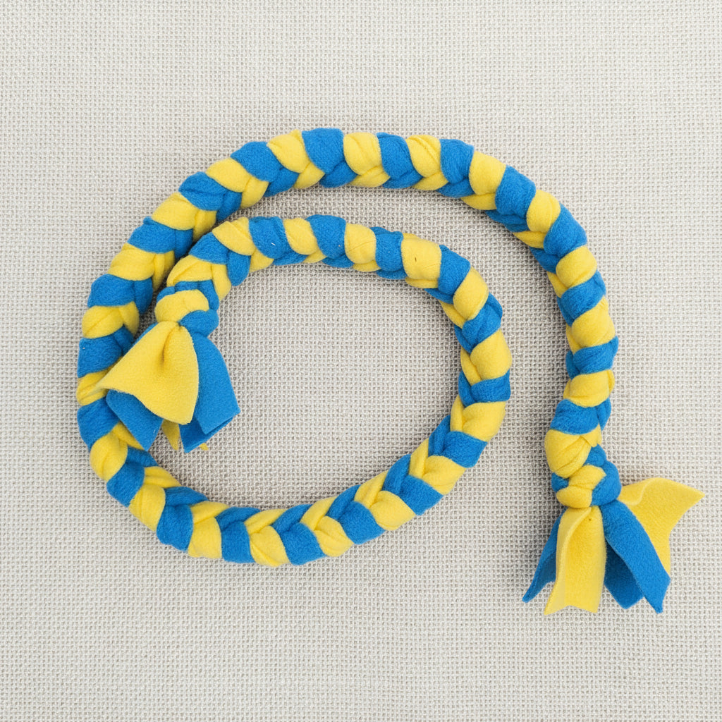 A braided rope dog toy in alternating blue and yellow stripes, laid out in a loop on a dark brown wooden surface. The toy features two small fabric bows at the ends, one in blue and one in yellow.