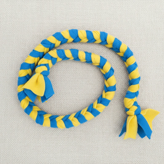 A braided rope dog toy in alternating blue and yellow stripes, laid out in a loop on a dark brown wooden surface. The toy features two small fabric bows at the ends, one in blue and one in yellow.