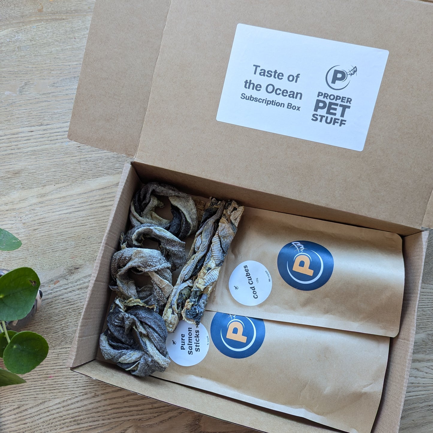 Our taste of the ocean subscription box is open showing the name on the open lid. You can see every item inside - the cod donuts and fish fingers are on the left of the box and the cod cubes and salmon stick are in brown craft bags on the right.