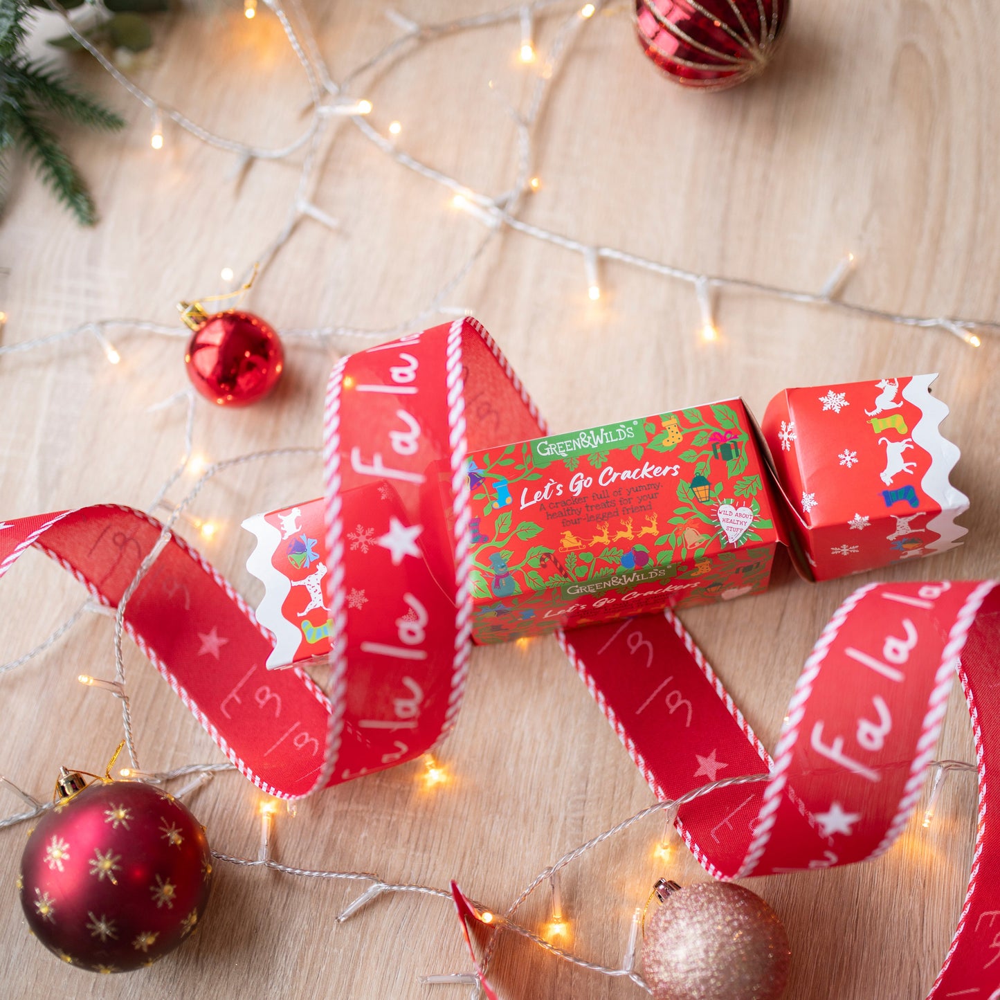 a festive print, red christmas cracker for dogs. It is made of cardboard, rectangular in shape. There is a white bauble at the bottom of the photo, a red one in the bottom right corner and a red and silver striped bauble at the top