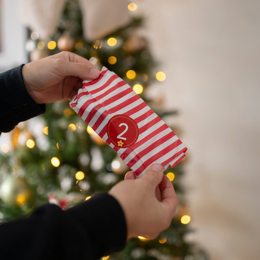 Red and white striped advent present with the number 2, held by a person in front of a Christmas tree.