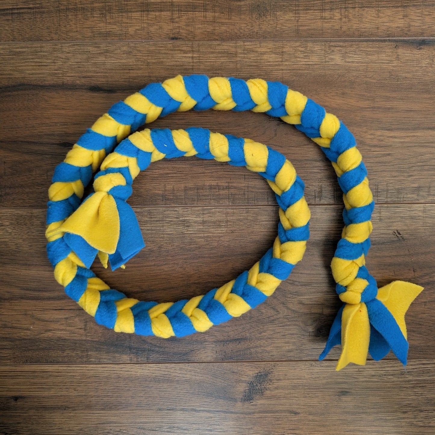 A braided rope dog toy in alternating blue and yellow stripes, laid out in a loop on a dark brown wooden surface. The toy features two small fabric bows at the ends, one in blue and one in yellow.