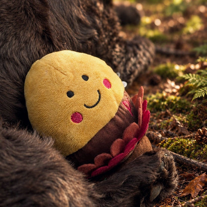 A plush toy shaped like a smiling yellow acorn with pink cheeks and a brown body, nestled against dark brown spaniel outdoors on a forest floor with moss and leaves.