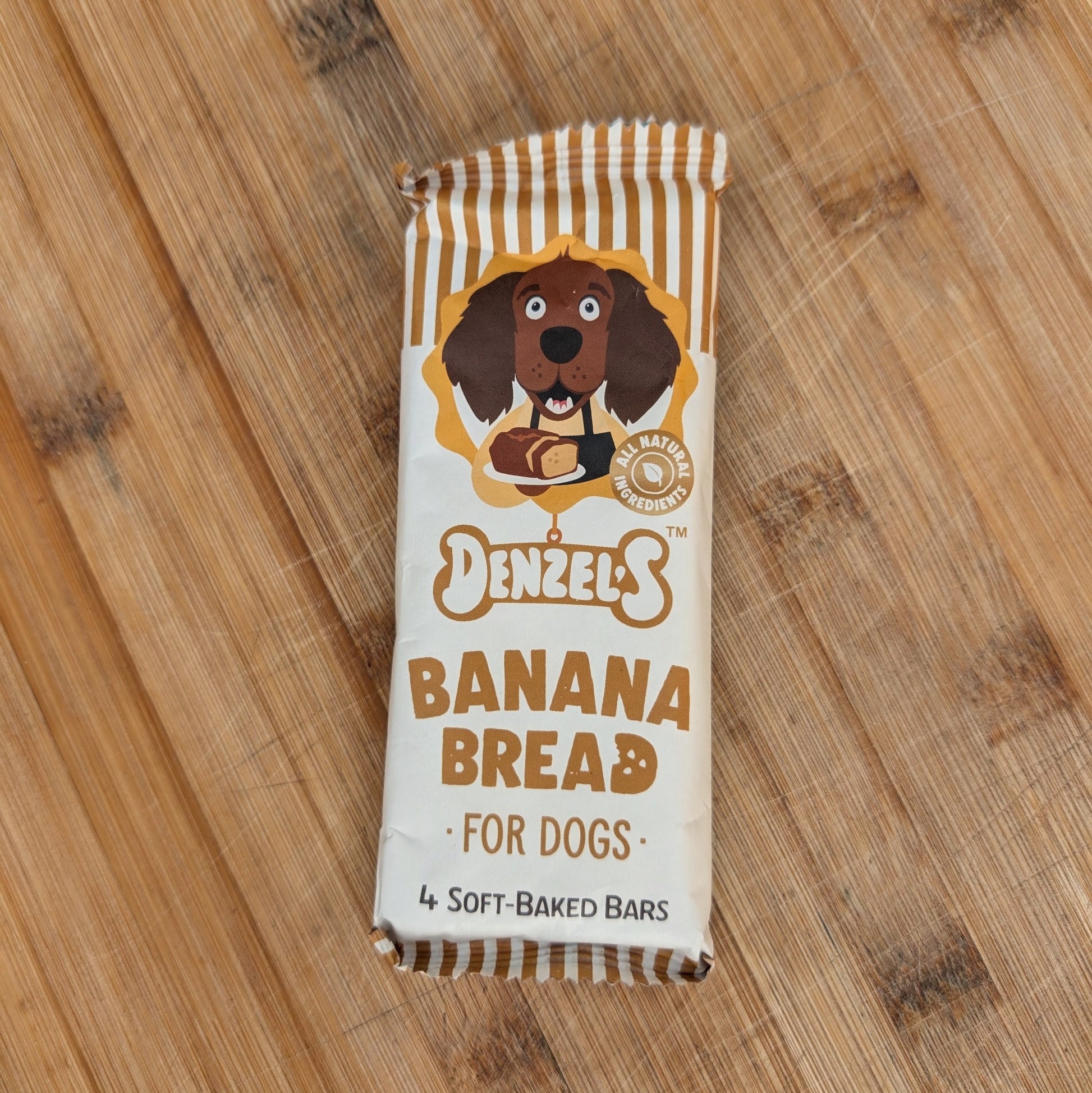 A wrapped dog banana bread bar from Denzels, featuring a cartoon dog illustration and brown text on a cream wrapper with white stripes, placed on a wooden surface.