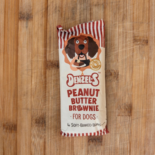 A package of Denzel's Peanut Butter Brownie for Dogs, featuring a cream-colored wrapper with red and white striped borders and an illustration of a dachshund, placed on a wooden surface.