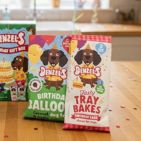 Two colorful Denzel's birthday dog treats on a wooden table in a kitchen setting. The front bag is for 'Tasty Tray Bakes Birthday Cake' coconut dog treats, with the Birthday Balloon Treats behind it