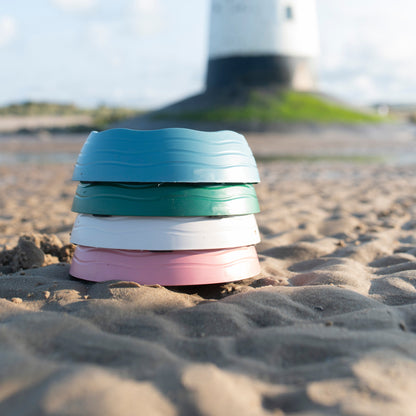 Stack of colorful dog bowls on a sandy beach with a lighthouse in the background