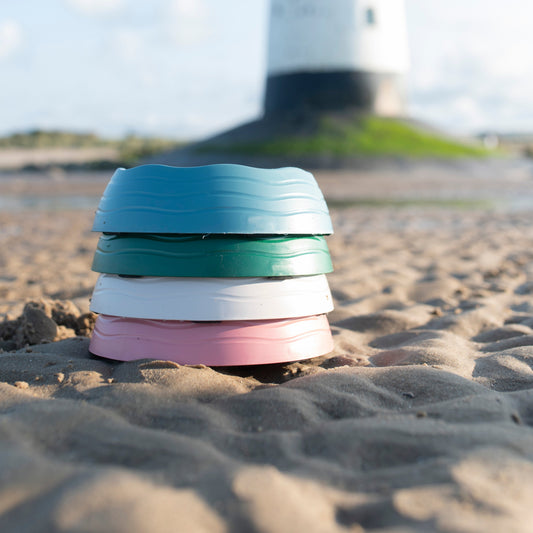 Stack of colorful dog bowls on a sandy beach with a lighthouse in the background