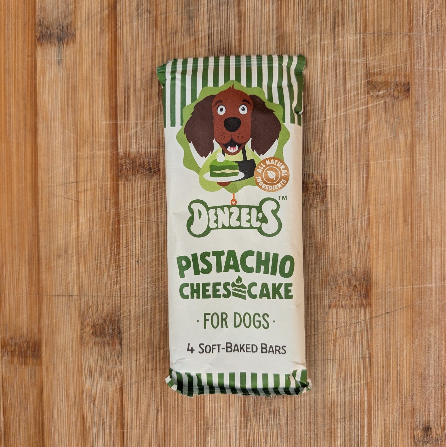 A package of Denzel's Pistachio Cheesecake dog treats, featuring a cream-colored wrapper with green stripes, a cartoon dachshund illustration, and green text, displayed on a wooden surface.