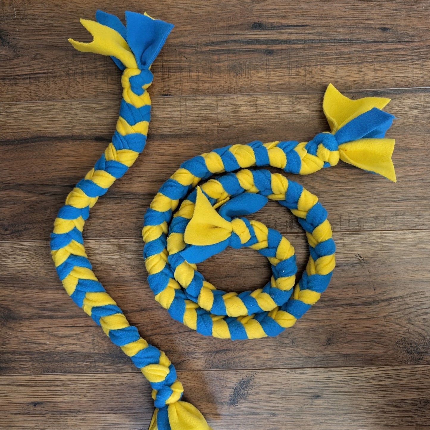 A braided dog toy made from blue and yellow fleece, arranged on a dark wood floor. The toy has a long braided body with blue and yellow alternating stripes and two knotted ends, one with a blue loop and yellow flaps, and the other with a yellow loop and blue flaps.
