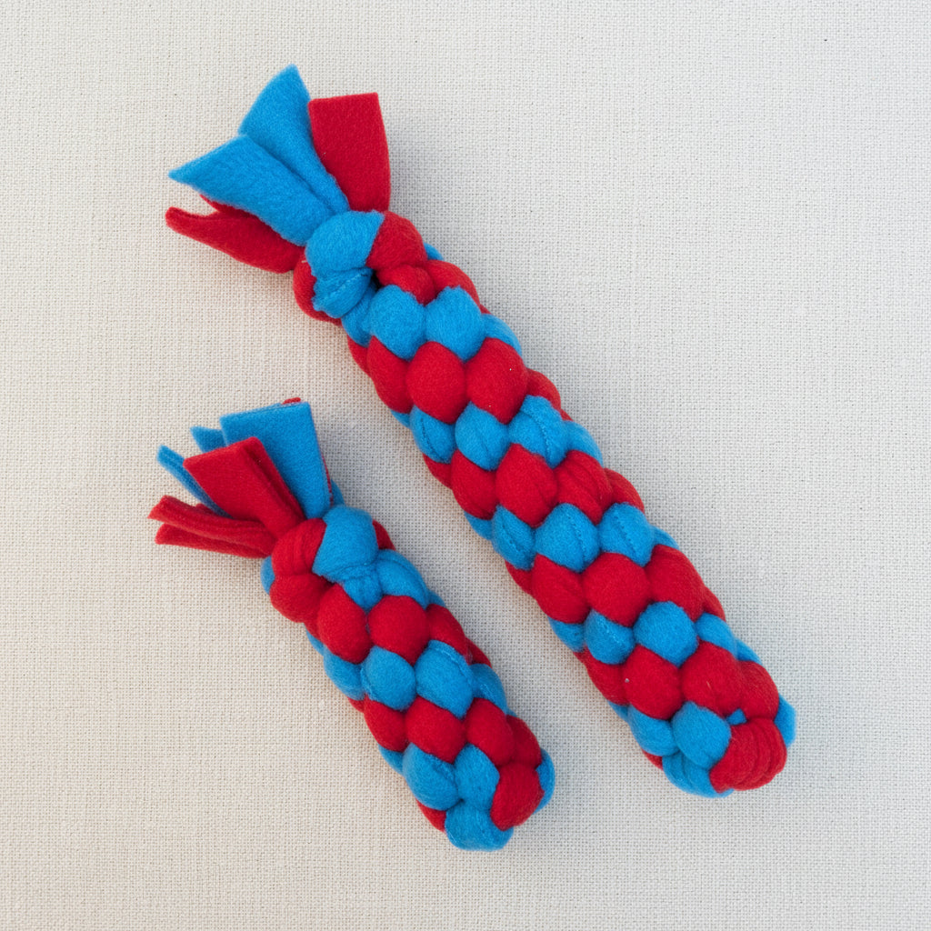 Two red and blue braided dog toys made from fleece with flared ends, displayed on a light beige textured background.
