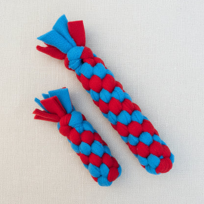 Two red and blue braided dog toys made from fleece with flared ends, displayed on a light beige textured background.