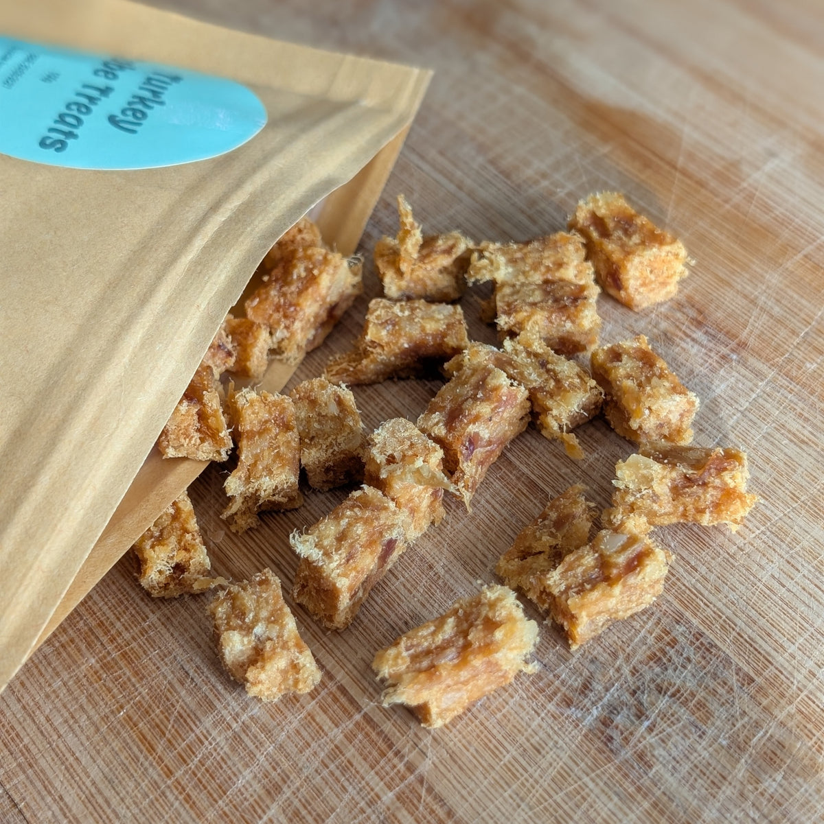 Turkey meat treats scattered on a wooden cutting board next to a brown paper pouch with a blue label.