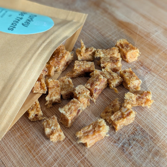 Turkey meat treats scattered on a wooden cutting board next to a brown paper pouch with a blue label.