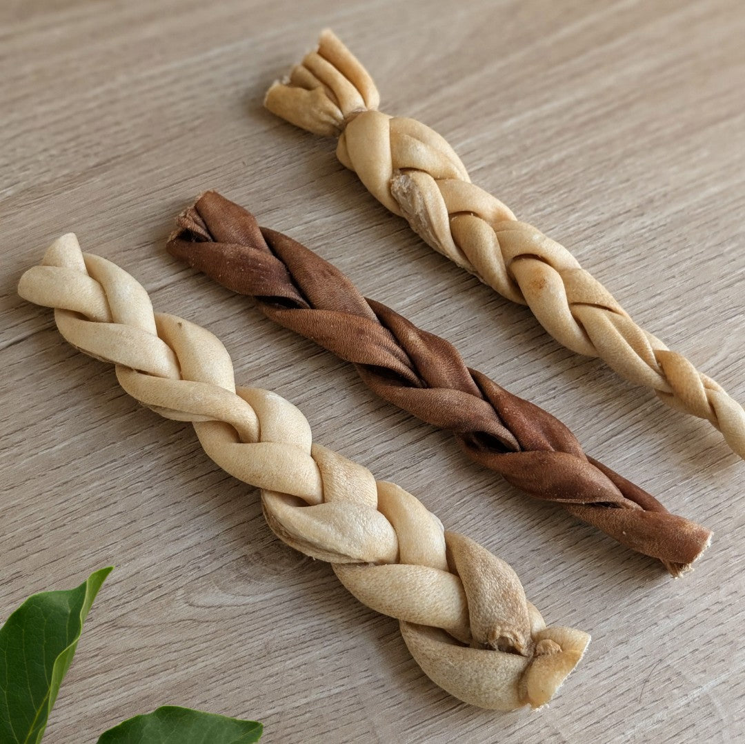 Three braided dog chew treats in light beige and medium brown, arranged diagonally on a light wood grain surface.
