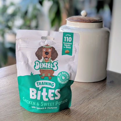 a green and white kraft bag of dog treats. There is a cartoon brown cocker spaniel on the bag. The bag is on the corner of a table in front of a white jar with a wooden lid. The jar is on the right behind the treats. There is a standing plant on the floor, blurred in the background in the top left corner