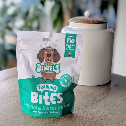 a green and white kraft bag of dog treats. There is a cartoon brown cocker spaniel on the bag. The bag is on the corner of a table in front of a white jar with a wooden lid. The jar is on the right behind the treats. There is a standing plant on the floor, blurred in the background in the top left corner