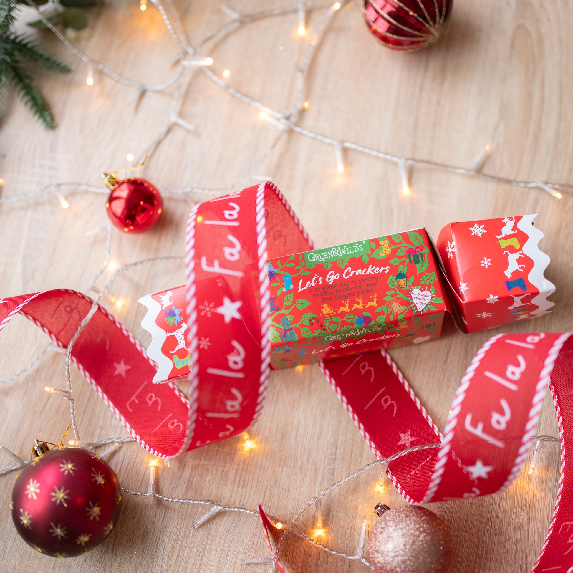 a festive print, red christmas cracker for dogs. It is made of cardboard, rectangular in shape. There is a white bauble at the bottom of the photo, a red one in the bottom right corner and a red and silver striped bauble at the top