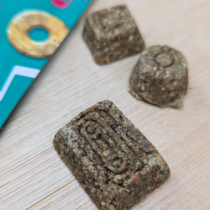 Three square-shaped denzels treats on a light wooden surface with a colorful book in the corner.