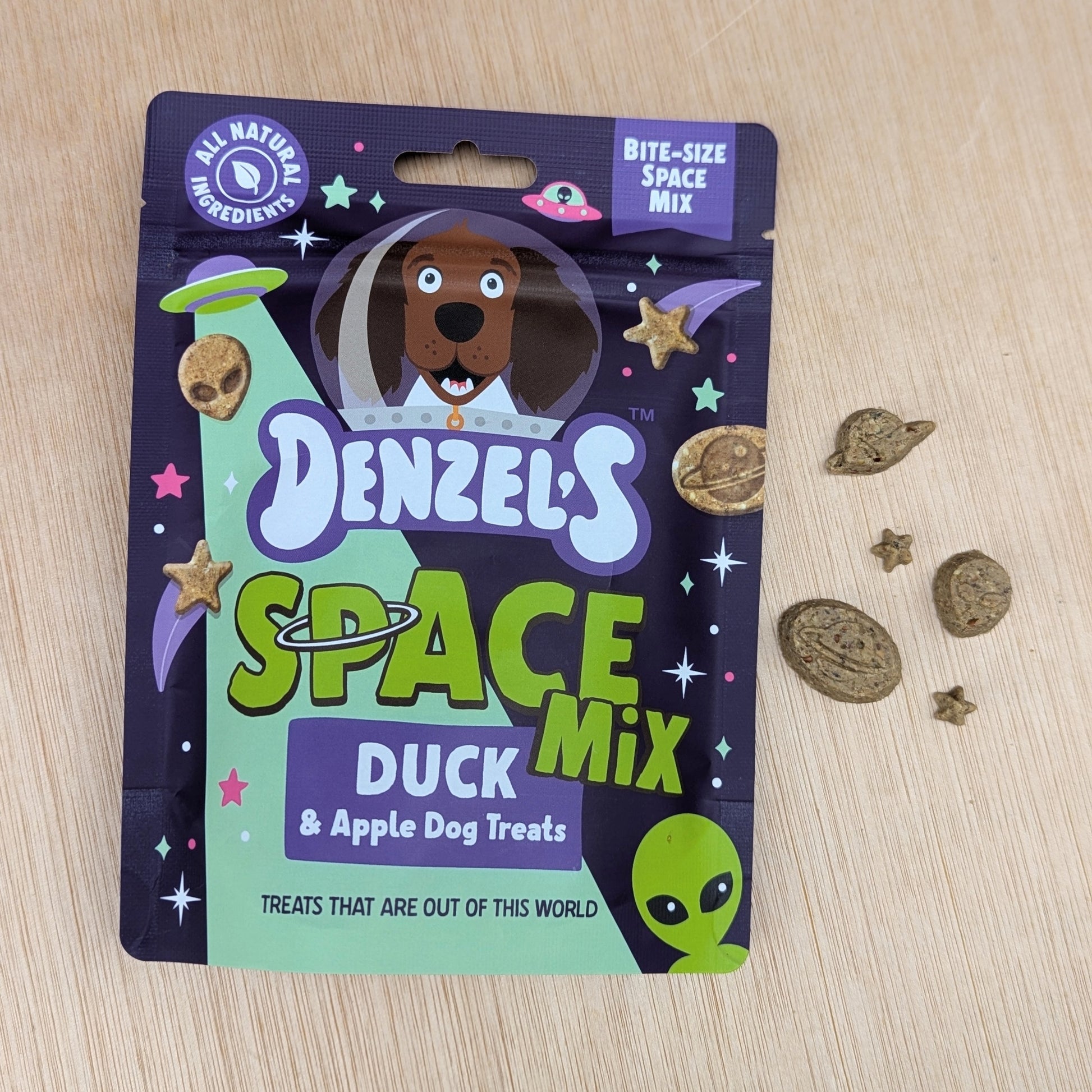 Denzel's Space Mix duck and apple dog treats packaging on a wooden surface with treats scattered on the right.
