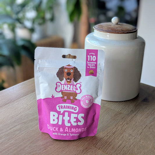 a pink and white kraft bag of dog treats. There is a cartoon brown cocker spaniel on the bag. The bag is on the corner of a table in front of a white jar with a wooden lid. The jar is on the right behind the treats. There is a standing plant on the floor, blurred in the background in the top left corner