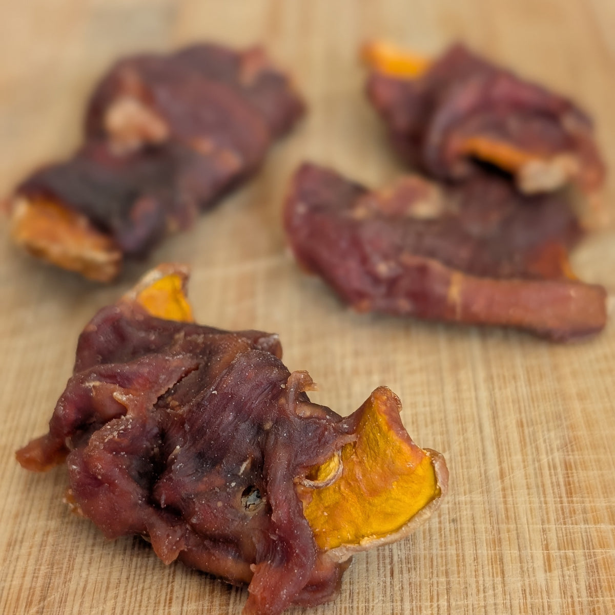 Four pieces of dried, dark reddish-brown duck meat wrapped around bright orange-yellow pimpkin, visible at the ends, arranged on a light wood cutting board.