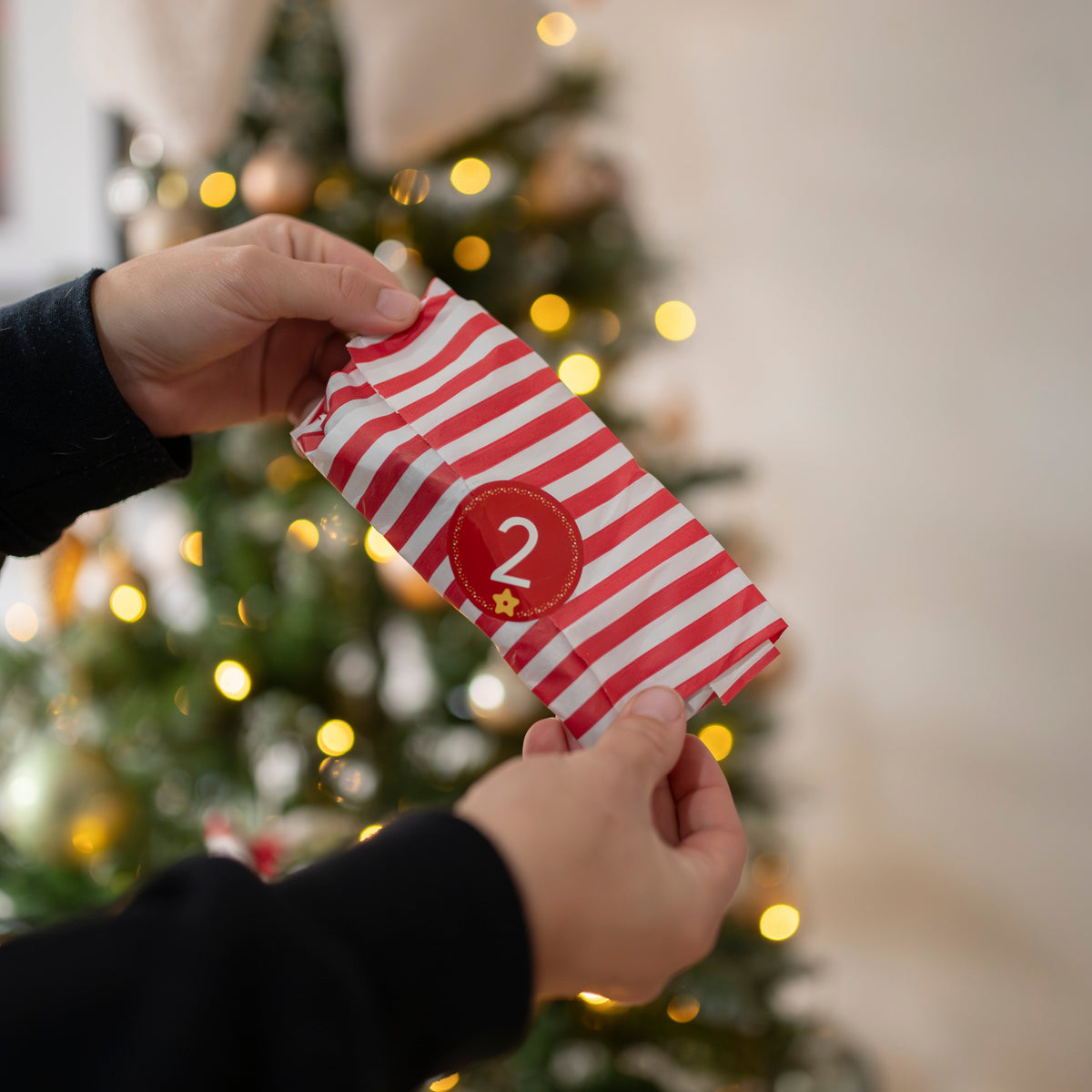 Red and white striped advent present with the number 2, held by a person in front of a Christmas tree.