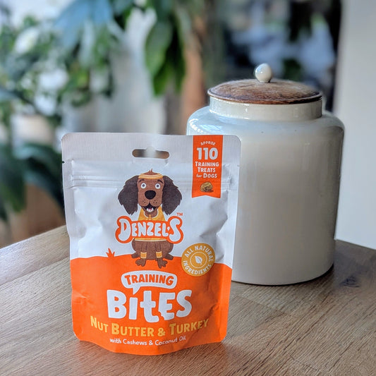 an orange and white kraft bag of dog treats. There is a cartoon brown cocker spaniel on the bag. The bag is on the corner of a table in front of a white jar with a wooden lid. The jar is on the right behind the treats. There is a standing plant on the floor, blurred in the background in the top left corner