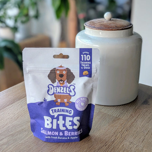 a blueish purple and white kraft bag of dog treats. There is a cartoon brown cocker spaniel on the bag. The bag is on the corner of a table in front of a white jar with a wooden lid. The jar is on the right behind the treats. There is a standing plant on the floor, blurred in the background in the top left corner