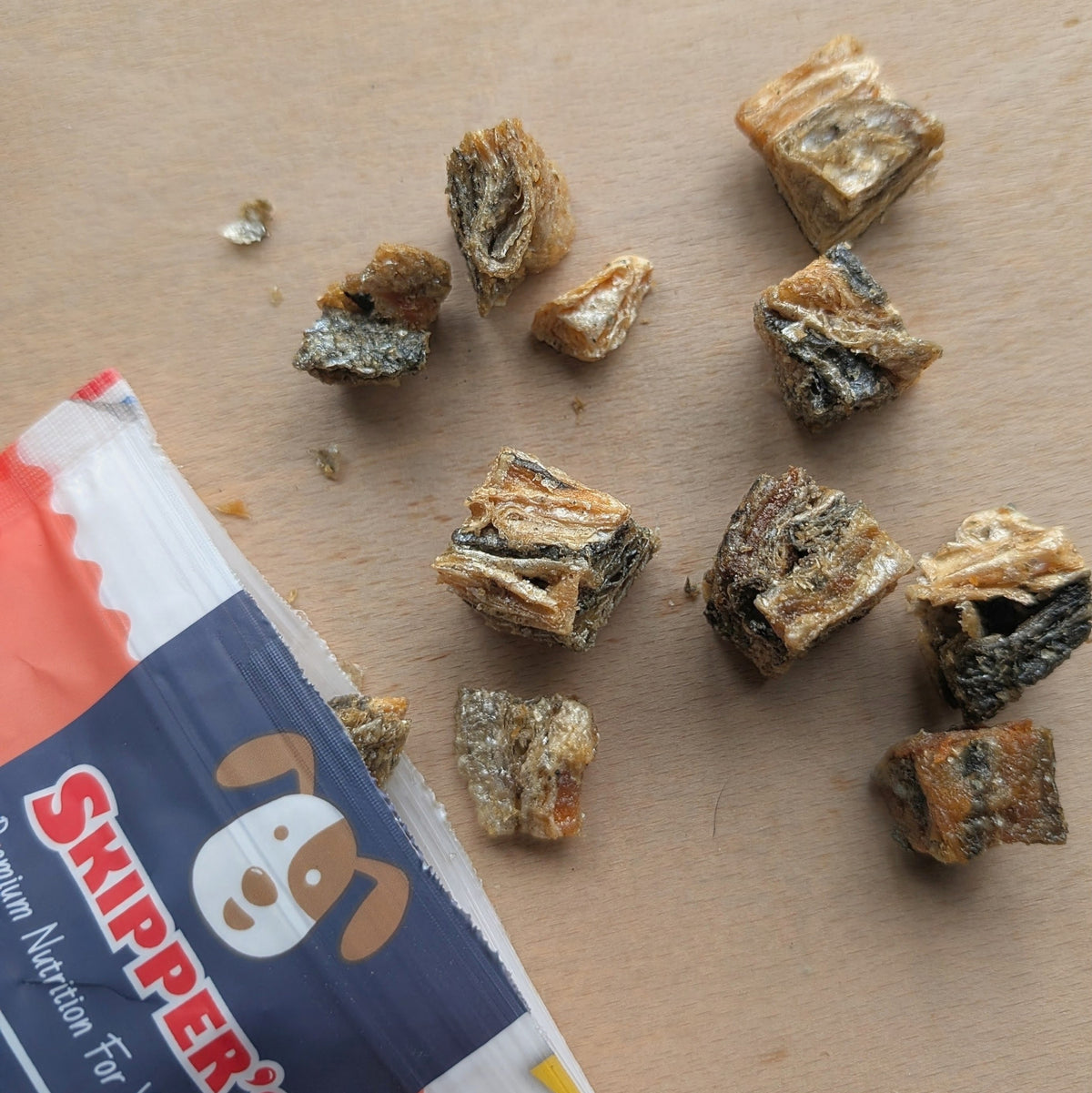 Several small pieces of dried salmon dog treats scattered on a light wood surface next to a partially visible Skipper's Premium Nutrition dog treat bag.