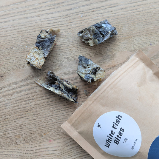 White fish skin treats spilling out of a brown bag. The treats are almost cube shape, made from fish skins. The brown bag is ziplock and a round white label stating the name can be seen.