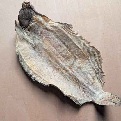A dried whole sole fish laid flat on a light beige surface, showing its natural pale tan and gray-brown skin with fine ribbed fins.